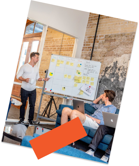 A man in front of a whiteboard showing a table to a man sitting on a sofa.