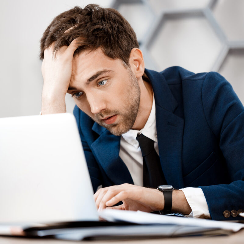 Upset young businessman sitting at laptop, office background.