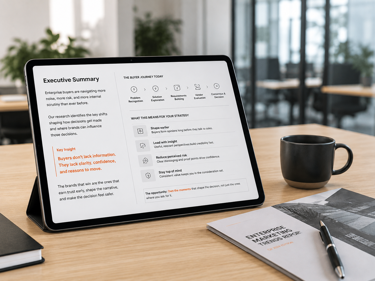 Alt text: Tablet on a light wood desk in a bright office, displaying an executive summary with buyer journey stages, strategy notes, and a highlighted insight about clarity, confidence, and decision-making, beside a report booklet, pen, and coffee mug.